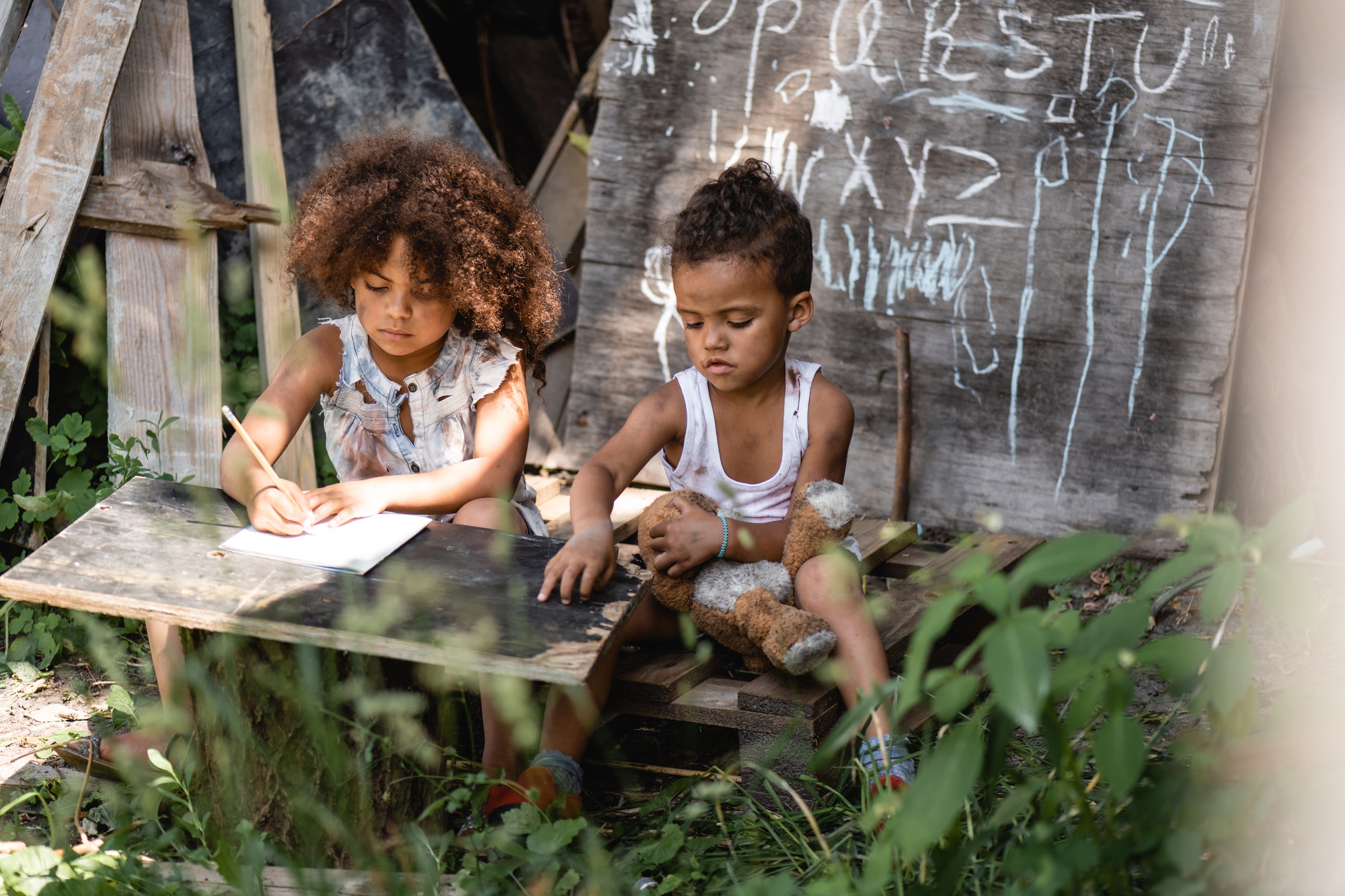selective focus of african american kid writing near brother sitting with dirty teddy bear
